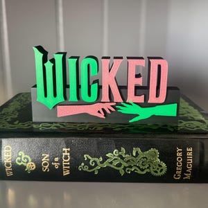 Könnte beinhalten: Ein grünes und pinkfarbenes Holzschild mit dem Wort "WICKED" in fetten Buchstaben. Das Schild liegt auf einem schwarzen Buch mit dem Titel "Wicked: Son of a Witch" von Gregory Maguire.