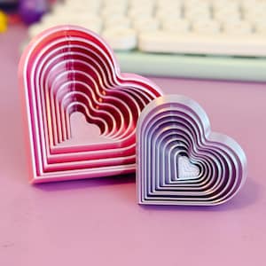 May include: Two heart-shaped cookie cutters in shades of pink and lavender. The cutters are nested, with a series of progressively smaller hearts inside each other. The larger cutter is a vibrant pink, while the smaller one is a soft lavender.