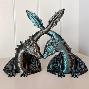 May include: Two dragon figurines, one black and one teal, face each other. Their tails cross in the background. The dragons have detailed scales, wings, and a spiky ridge down their backs. The figurines are on a light-colored surface.