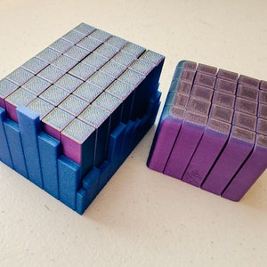 May include: Two sets of colorful magnetic building blocks. One set is a large rectangular prism with a blue exterior and a purple interior. The other set is a smaller cube with a purple exterior and a blue interior.