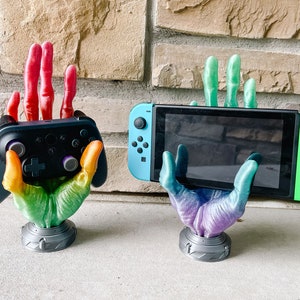 Könnte beinhalten: Zwei 3D-gedruckte Hände halten einen Videospiel-Controller und eine Nintendo Switch-Konsole. Die Hände sind in Regenbogenfarben bemalt. Der Controller ist schwarz mit violetten Knöpfen. Die Switch-Konsole ist blau und grün.
