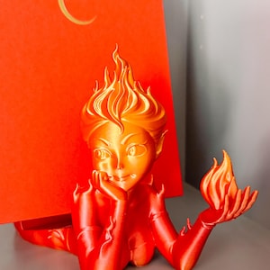 May include: A 3D-printed figurine of a female figure with fiery hair and a flame in her hand, in shades of orange and red. The figure is posed in front of a red background with a crescent moon design.
