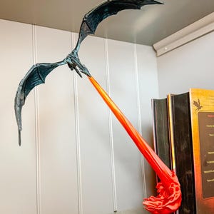 May include: A black dragon figurine with outstretched wings, breathing a bright orange and red flame. The dragon is perched on a bookshelf with several books behind it.