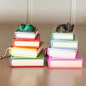 May include: Two stacks of colorful miniature books with white pages and a gold chain. One stack has a green and red sculpture on top, the other has a dark cat sculpture. The books are in shades of red, orange, yellow, green, blue, and pink.