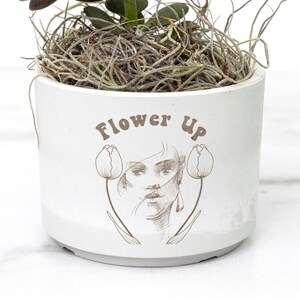 May include: A white ceramic planter with the text "Flower Up" and a line drawing of a woman's face with two tulips. A small green succulent plant is growing in the planter.