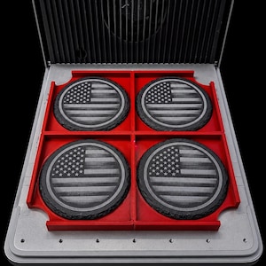 May include: Four circular black slate coasters featuring an engraved American flag design. The coasters are arranged in a red plastic grid holder with a silver metallic base and a black vertical slatted panel in the background.
