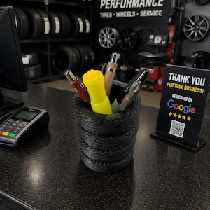 May include: A black cylindrical pencil holder designed to resemble a stack of tires, containing several pens and a bright yellow tool. The holder sits on a dark countertop next to a payment terminal and a sign that reads THANK YOU FOR YOUR BUSINESS.