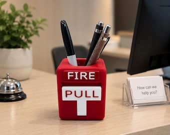Op de afbeelding: Een rode pennenhouder in de vorm van een brandmeldcentrale, met de witte woorden "FIRE PULL". Hij bevat een zwarte stift en twee pennen. De achtergrond toont een bureau, een computerscherm en een bord met "Welcome".