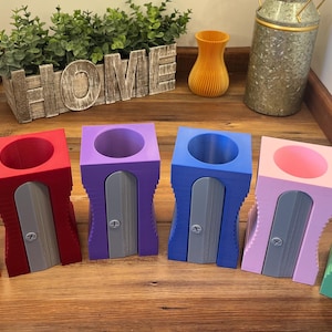 May include: Seven colorful pencil sharpeners with a gray metal blade. The sharpeners are arranged in a row on a wooden surface. The colors are orange, red, purple, blue, pink, and green.