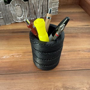 May include: A black pen holder with a tire tread design, filled with pens and markers. The pens include a red pen, a yellow highlighter, a wooden pen, and a black marker. The holder sits on a wooden surface.