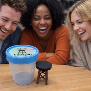 May include: A clear plastic container with a blue lid labeled "Stool Sample" sits on a wooden table next to a miniature stool. Three people are smiling and laughing in the background. The miniature stool is brown with a black seat.