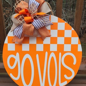 May include: Orange and white checkered round door hanger with the words "go vols" in white lettering. The door hanger is decorated with a large orange, white, and brown bow.