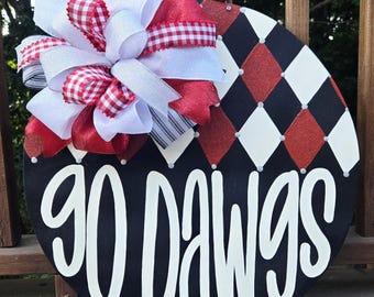 May include: A round wooden door sign featuring a black, white, and red diamond pattern on the upper half and the text "GO DAWGS" in large white script on the lower half. A large decorative bow with red, white, and gingham ribbons is attached.