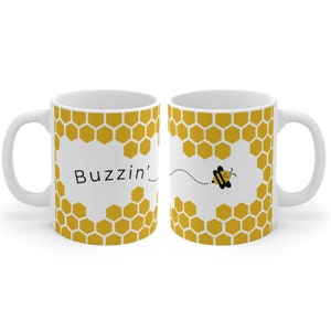 Puede incluir: Taza de cerámica blanca con un patrón de panal de abeja amarillo y una abeja de dibujos animados que sale del panal. La taza dice "Buzzin'" en el lado izquierdo.