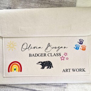 May include: A white felt folder with the text "Olivia Bouzan Badger Class" and "Art Work". The folder has a rainbow, a sun, stars, a badger, and handprints.