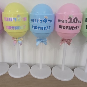 May include: Five colourful lollipop-shaped birthday decorations. Each has a different colour, including purple, yellow, blue, pink, and green. Each lollipop has a name and age printed on it, with a bow at the base. The decorations are on white stands.