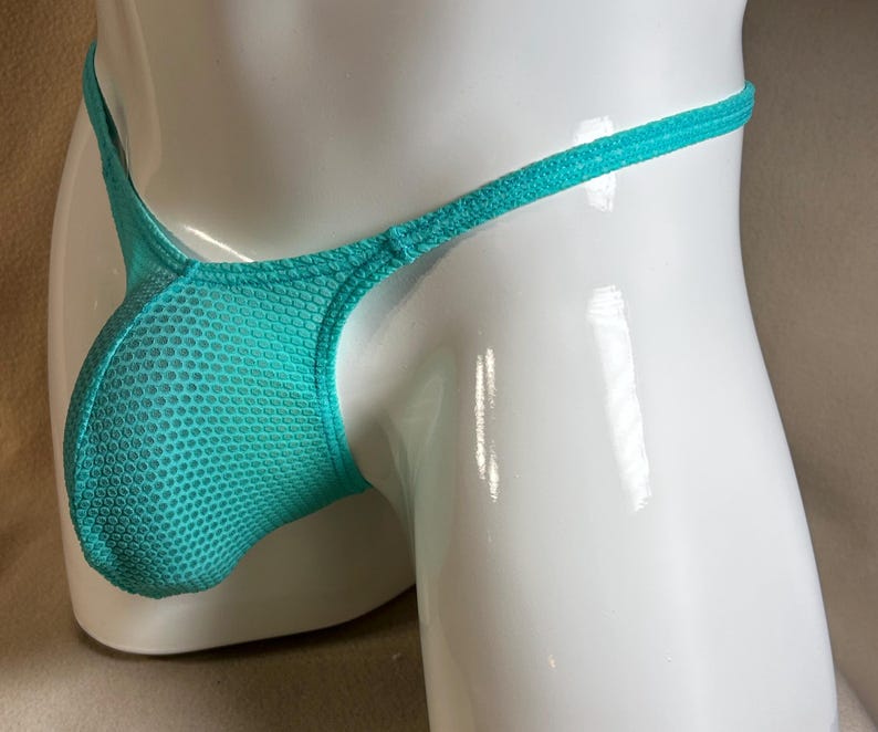May include: A turquoise thong made of a mesh fabric. The garment has a pouch in the front and thin straps. The thong is displayed on a white mannequin.