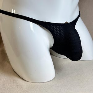 May include: Black thong underwear with a mesh pouch and thin straps. The underwear is displayed on a white mannequin. The fabric appears to be a textured knit.