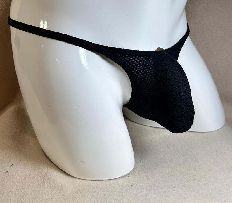 May include: Black thong underwear with a mesh pouch and thin straps. The underwear is displayed on a white mannequin. The fabric appears to be a textured knit.