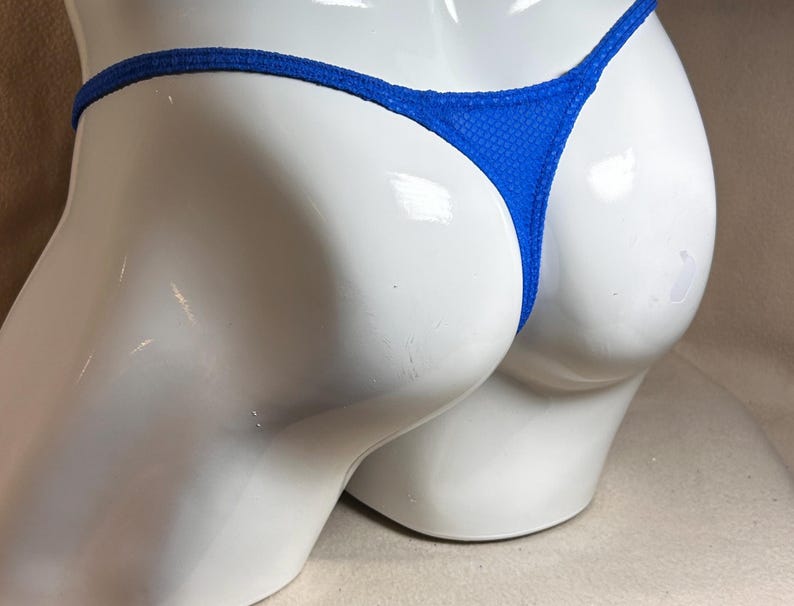 May include: A close-up of a blue thong swimsuit. The swimsuit is displayed on a white mannequin. The thong has a textured pattern and a thin waistband.