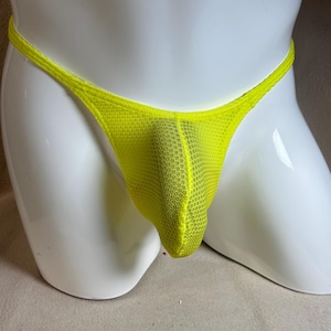 May include: A neon yellow thong underwear made of a mesh fabric. The underwear is displayed on a white mannequin. The thong features a thin waistband and a pouch in the front.