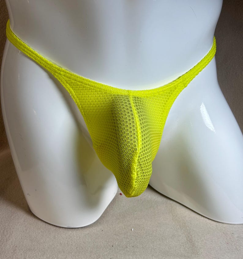 May include: A neon yellow thong underwear made of a mesh fabric. The underwear is displayed on a white mannequin. The thong features a thin waistband and a pouch in the front.