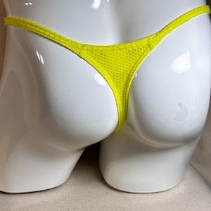 May include: Bright yellow thong underwear made of a mesh fabric. The garment is displayed on a white mannequin. The thong has a thin waistband and a minimal back coverage.