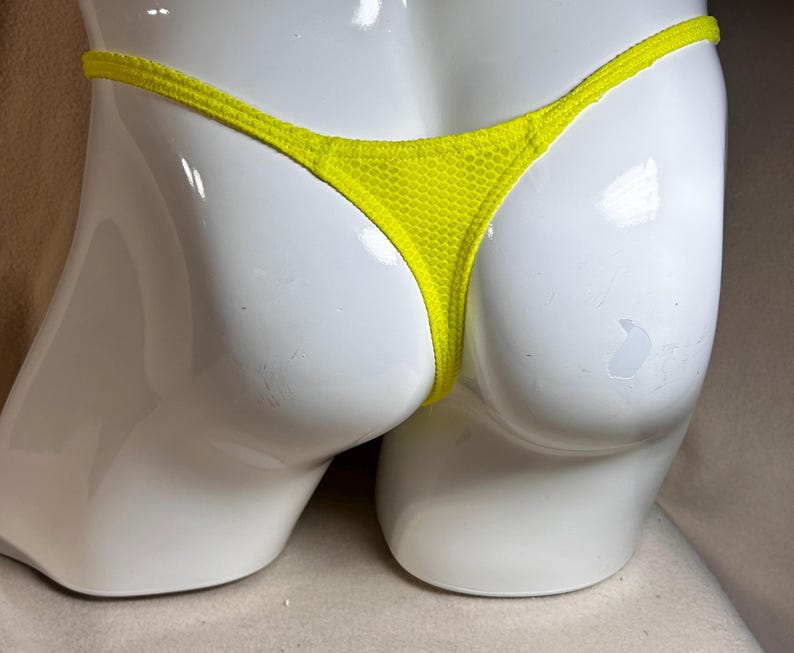 May include: Bright yellow thong underwear made of a mesh fabric. The garment is displayed on a white mannequin. The thong has a thin waistband and a minimal back coverage.