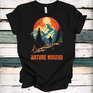 May include: Black t-shirt featuring a retro-style graphic of a mountain range with a person standing on a peak. The graphic has a sunset background with a yellow moon and birds flying. The text "NATURE NOMAD" is printed below the graphic.