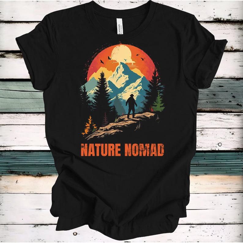 May include: Black t-shirt featuring a retro-style graphic of a mountain range with a person standing on a peak. The graphic has a sunset background with a yellow moon and birds flying. The text "NATURE NOMAD" is printed below the graphic.