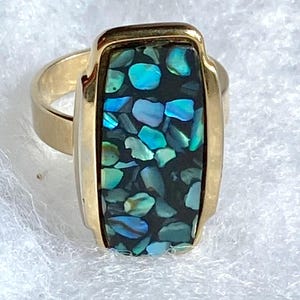 May include: A gold ring with a rectangular abalone shell inlay. The shell is a mosaic of blue, green, and black iridescent pieces.
