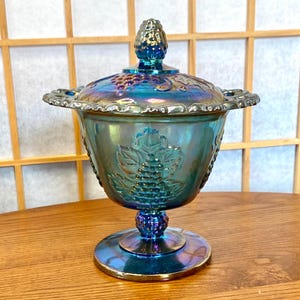 May include: An iridescent blue glass candy dish with a lid. The dish features a grape design on the bowl and lid. The dish has a pedestal base and a decorative rim. The lid has a small knob on top.