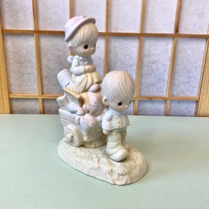 May include: A ceramic figurine featuring two children. One child is seated on a wagon, wearing a hat and dress. The other child stands nearby. The figurine is primarily cream-colored with pastel accents and sits on a base resembling grass.