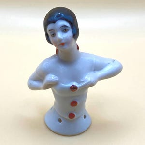 May include: A vintage porcelain half-doll figurine. The doll has a white body, dark blue hair, and red accents on the buttons and earrings. The doll's arms are raised, and it has a painted face with red lips.