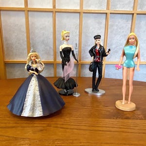 May include: Four collectible dolls standing in a row. The first wears a dark blue and silver gown with a crown. The second wears a black strapless dress with a pink scarf and stands by a microphone. The third wears a black leather jacket and pants. The fourth wears a light blue swimsuit.