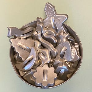 May include: A collection of various silver-toned metal cookie cutters packed inside a round metal container. The assortment includes shapes such as a gingerbread person, a star, a crescent moon, a flower, and other assorted holiday-themed designs.