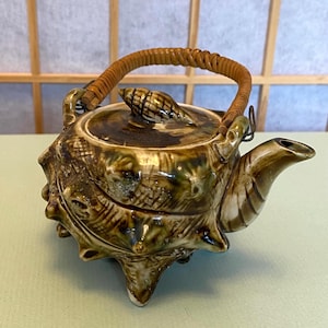 May include: A brown and green ceramic teapot with a shell design and a woven bamboo handle. The teapot has a spout and a lid with a shell knob.
