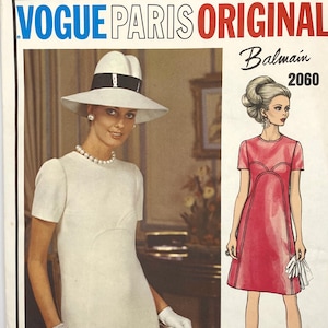 May include: A vintage Vogue Paris Original sewing pattern featuring a dress design by Balmain. The pattern number is 2060. The pattern shows a white dress with a scoop neck and short sleeves, and a pink dress with a scoop neck and short sleeves. Both dresses have a fitted bodice and a slightly flared skirt.