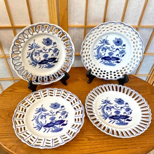 May include: Four white porcelain plates with blue floral designs. Each plate has a unique openwork or scalloped edge. The plates are displayed on black stands, showcasing their intricate patterns and craftsmanship.