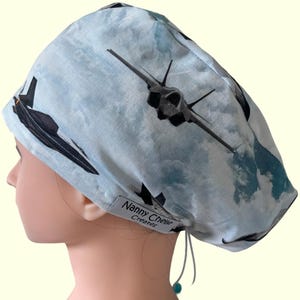 May include: A blue and white scrub hat with a print of fighter jets in the clouds. The hat has a white drawstring and a black label that reads "Nanny Chelle Creates".