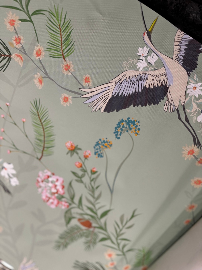 May include: A green background with a white crane with black wings and a yellow beak flying over a floral pattern. The flowers are pink, white, and blue.