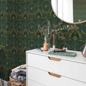Op de afbeelding: Een witte commode met drie laden en houten handgrepen. De commode heeft gouden metalen poten en staat voor een muur met een groen en gouden geometrisch patroon behang. Een ronde spiegel met een gouden lijst hangt aan de muur boven de commode.