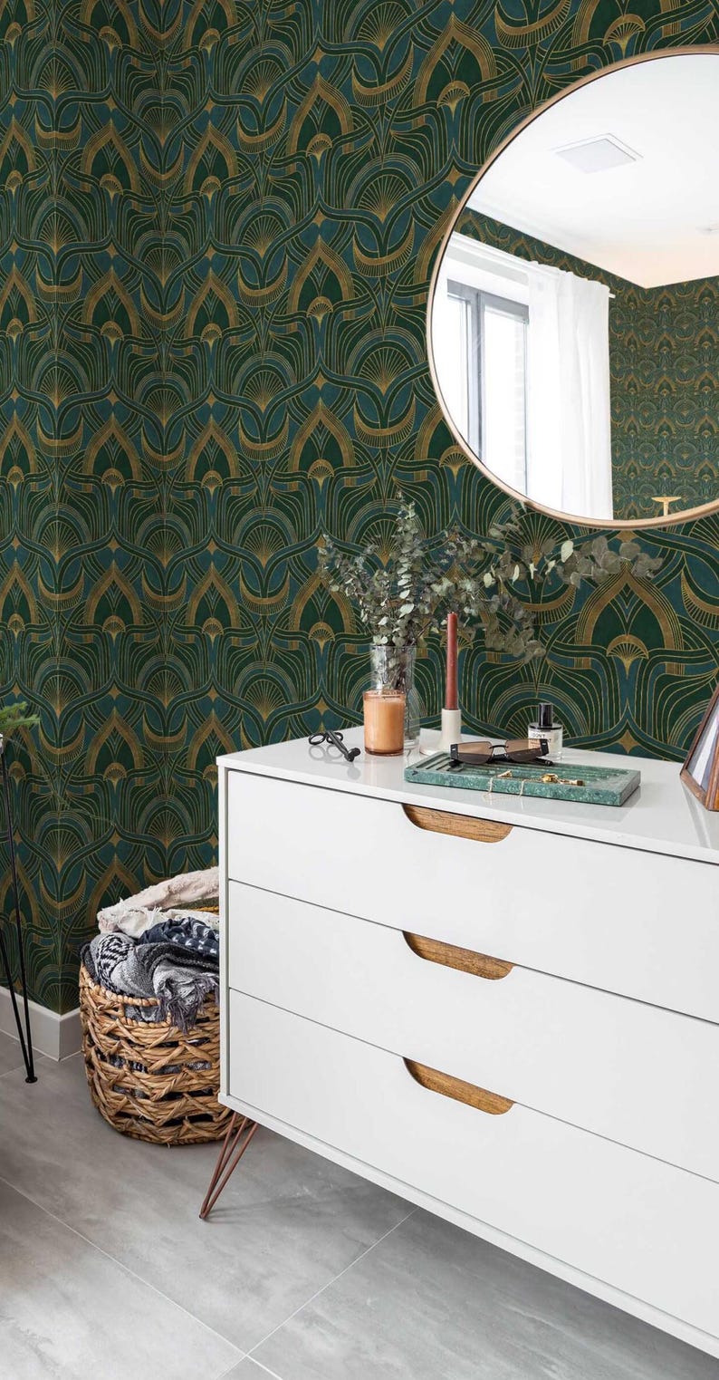 Op de afbeelding: Een witte commode met drie laden en houten handgrepen. De commode heeft gouden metalen poten en staat voor een muur met een groen en gouden geometrisch patroon behang. Een ronde spiegel met een gouden lijst hangt aan de muur boven de commode.