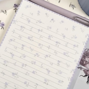 Könnte beinhalten: Ein liniertes Notizbuch mit floralem Muster und einem lila Stift. Das Notizbuch hat eine zarte Spitzenborte und ist mit kleinen lila Blumen bedeckt. Der Stift liegt obenauf und ist einsatzbereit.