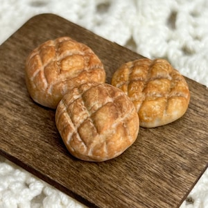 May include: Three miniature, golden-brown bread rolls with a textured, cross-hatched surface, presented on a small, dark wooden cutting board. The rolls have a slightly uneven shape, suggesting a homemade quality. The background is a soft, white, crocheted fabric.