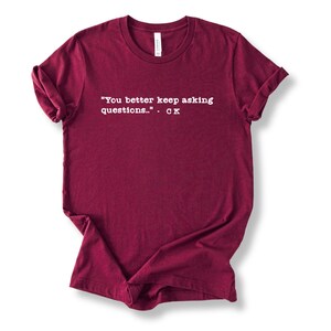 May include: A maroon t-shirt with rolled-up sleeves. The shirt features white text that reads: "You better keep asking questions..." - CK. The shirt is made of a soft material.