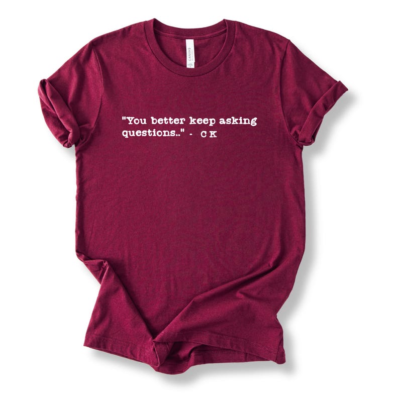 May include: A maroon t-shirt with rolled-up sleeves. The shirt features white text that reads: "You better keep asking questions..." - CK. The shirt is made of a soft material.