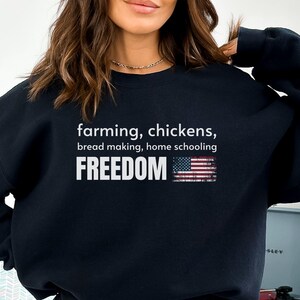 Può includere: Felpa nera con la parola "FREEDOM" in lettere bianche e in grassetto, insieme alle frasi "farming, chickens, bread making, home schooling". Una grafica della bandiera americana è a destra della parola "FREEDOM".