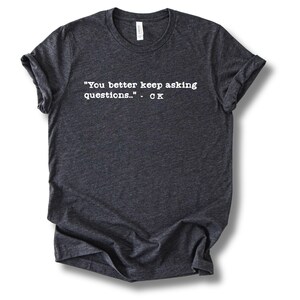 May include: Dark gray t-shirt with rolled-up sleeves. The shirt features white text that reads: "You better keep asking questions.." - CK. The shirt is made of a soft material and has a crew neck.