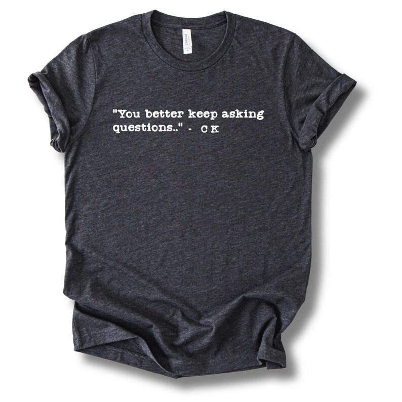 May include: Dark gray t-shirt with rolled-up sleeves. The shirt features white text that reads: "You better keep asking questions.." - CK. The shirt is made of a soft material and has a crew neck.